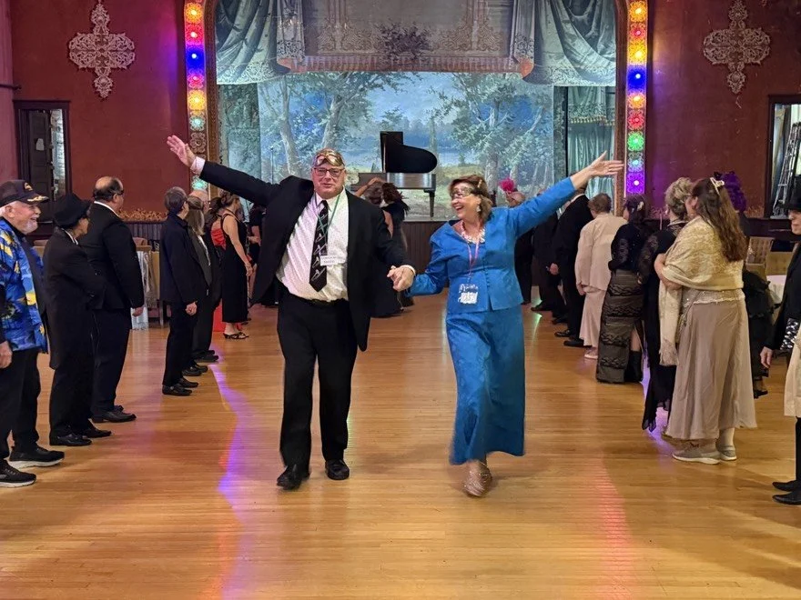 A group of people at a Masquerade Ball in formal attire dancing in a decorated hall with a wooden floor, colorful lights, and a backdrop of a scenic landscape with trees and water.