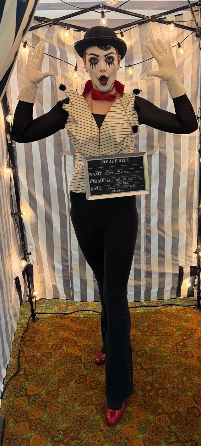 The mime of the circus poses with hands raised inside a circus tent decorated with string lights, holding a mock police department sign wearing clown makeup, a black bowler hat, striped vest, black pants, red shoes, and white gloves, 