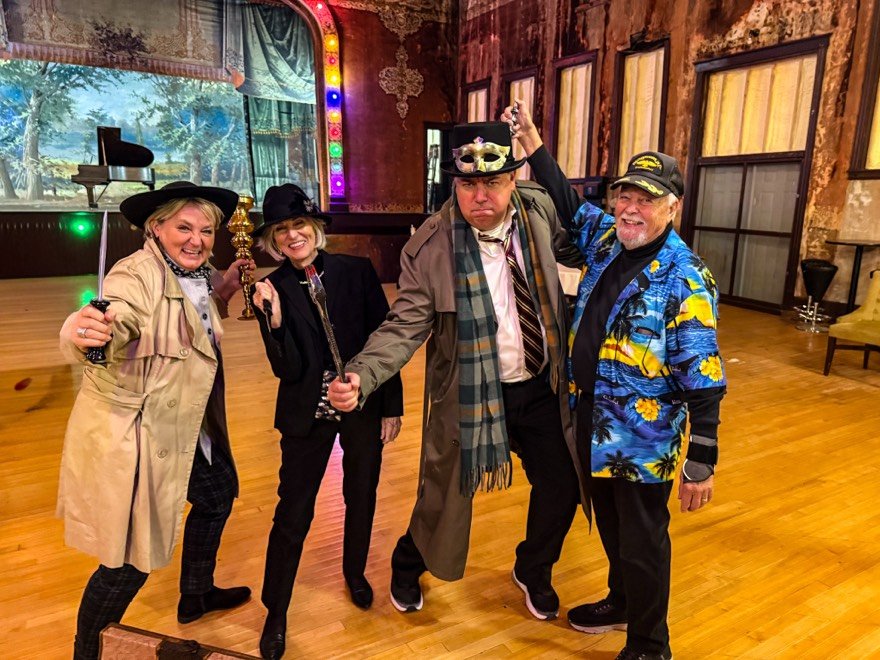 Four adults in detective costumes posing with fake murder weapons in a decorated room with wooden floors and walls, with a stage holding a baby grand piano in the background.