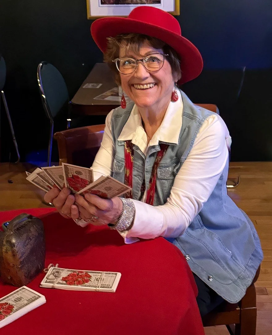 A smiling woman wearing a wide brim red hat, glasses, and a denim vest sitting at a table, holding fake money in her hands.
