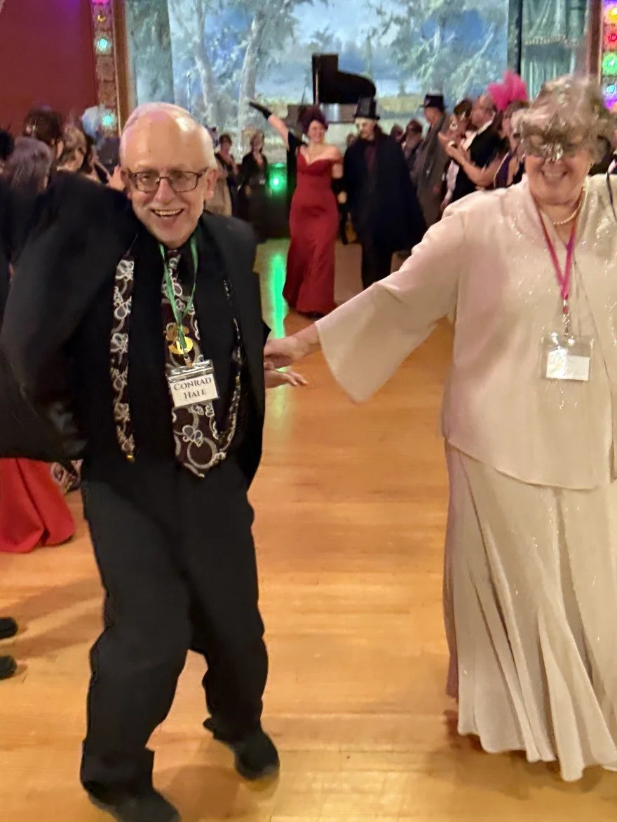 People dancing and enjoying themselves at the Phantom of the Batcher Block Opera House Masquerade Ball. Conrad, the lawyer, is wearing a suit and glasses smiling whole holding hands with Isla, a woman in a beige dress with a hat, both wearing name ta