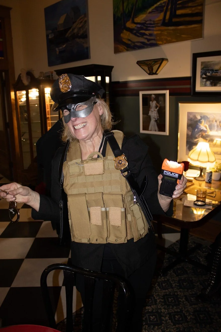 Skyler Peeks is dressed as a police officer with a mask, police hat, and tactical vest, smiling and holding police props in a warmly lit, decorated room.