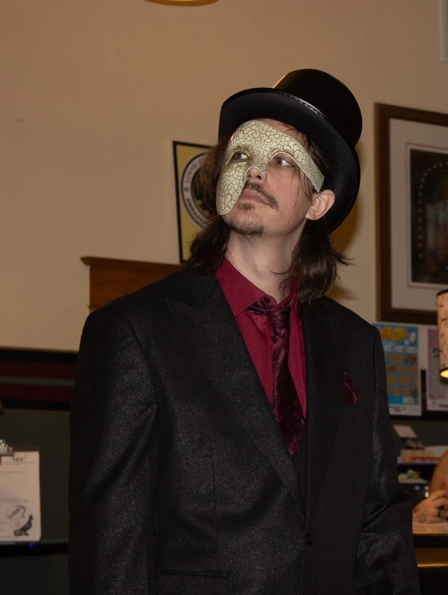 The Phantom of the Batcher Block Opera House, dressed in a black suit with a dark red shirt and tie is wearing a black top hat and an eye mask resembling a cracked desert landscape, looking slightly upward.