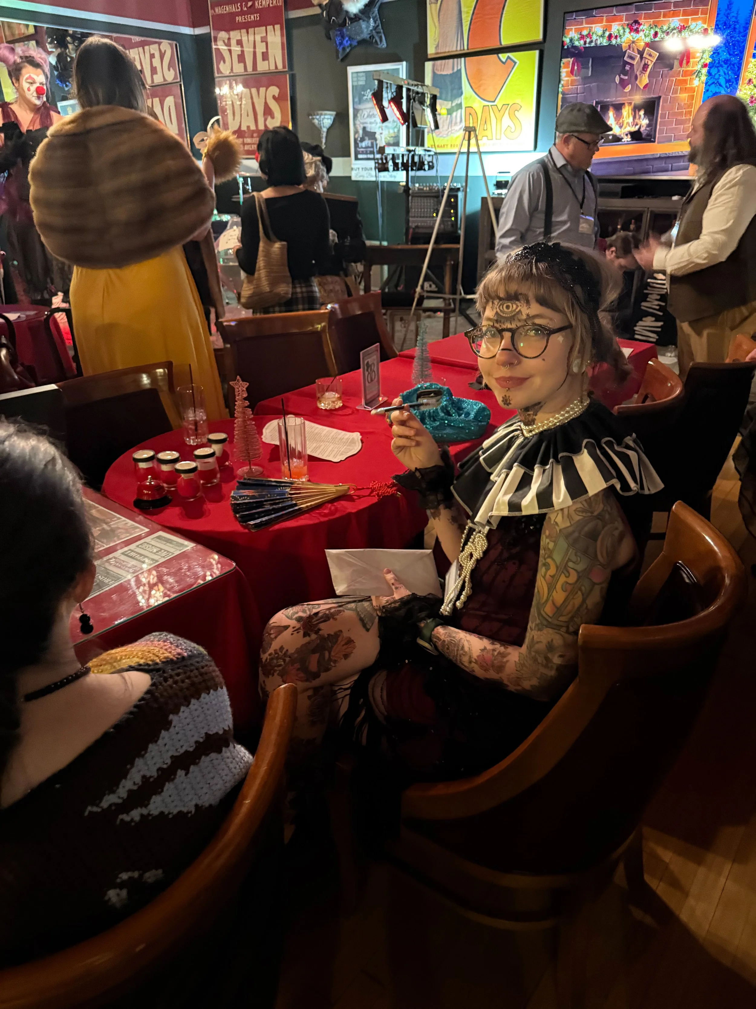 The most  tattooed person is dressed in a vintage style with a ruffled collar, pearls, and gloves, holding a cigarette holder. She sits at a table with a puzzle on it. 