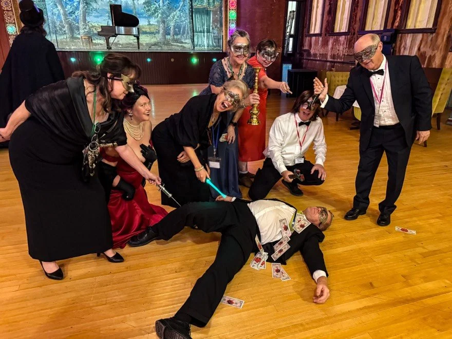 Group of people dressed in formal attire with masks, engaging in playful pretend crime scene with fake weapons at a Murder Mystery Party in a ballroom. The Murder Mystryss is kneeling near the assumed victim who is covered in fake money.