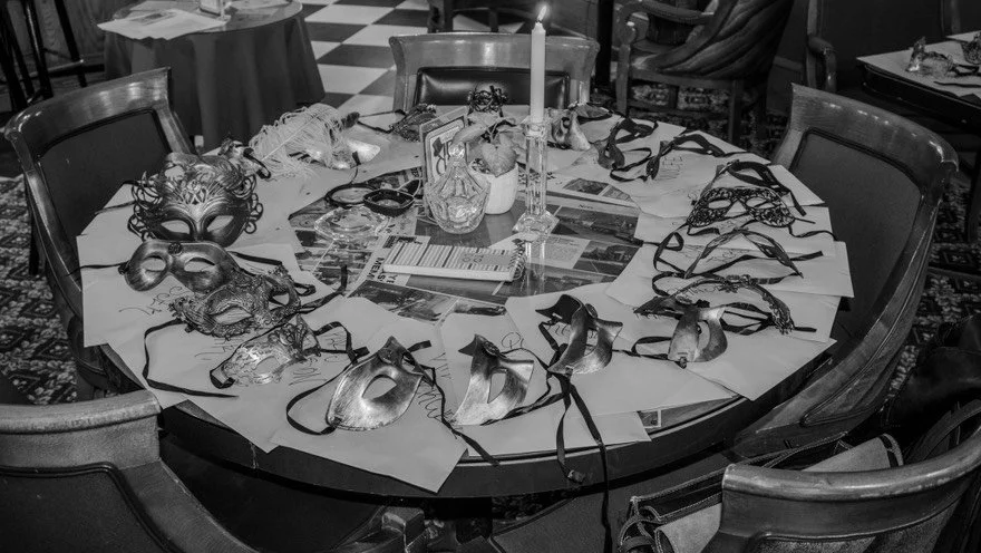 A greyscale image of a round table covered with various masks, papers, and decorative items, with chairs around it in a dining room setting.