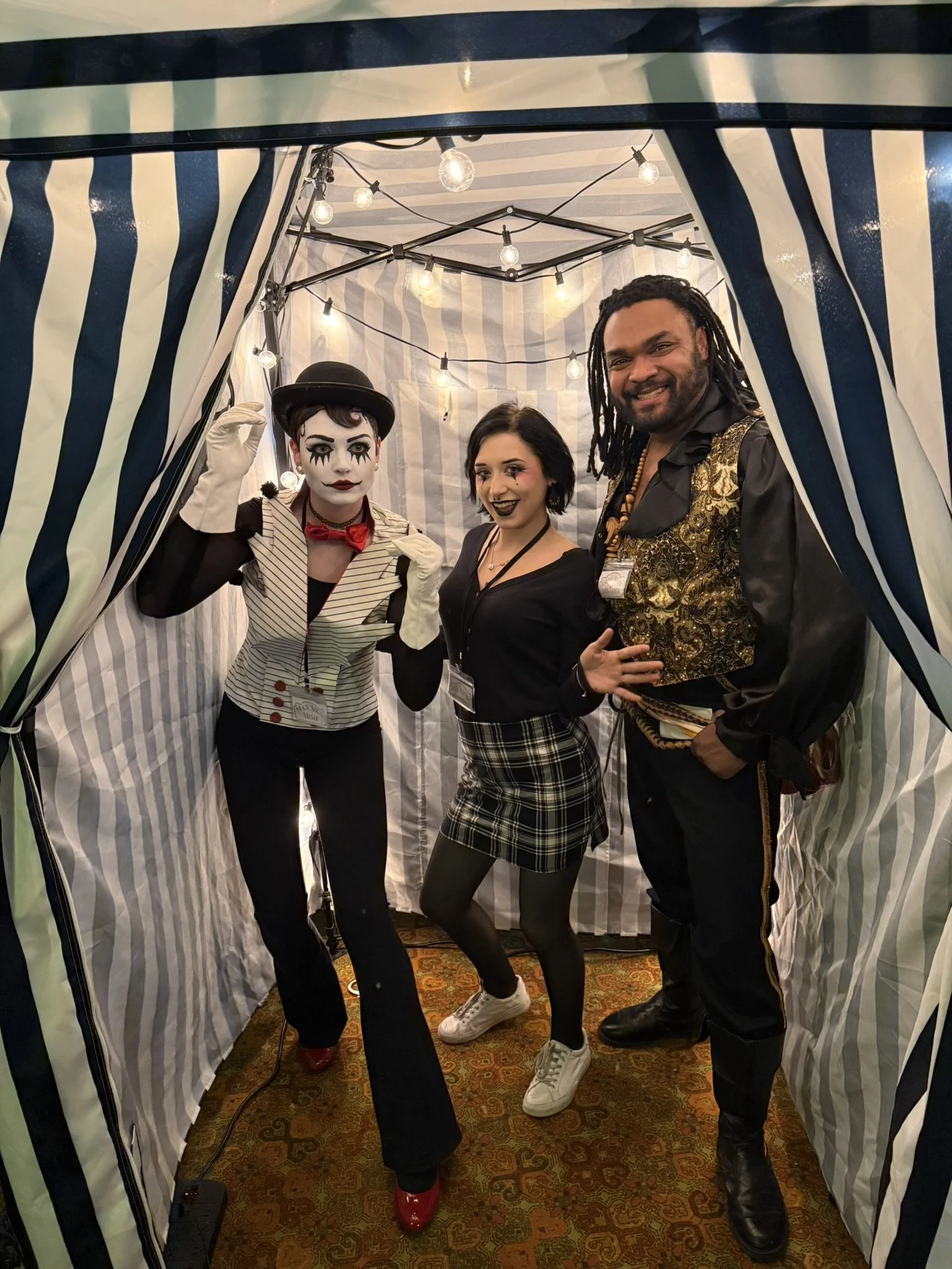 The mime, contortionist, and lion tamer posing together in a circus tent with string lights hanging overhead.