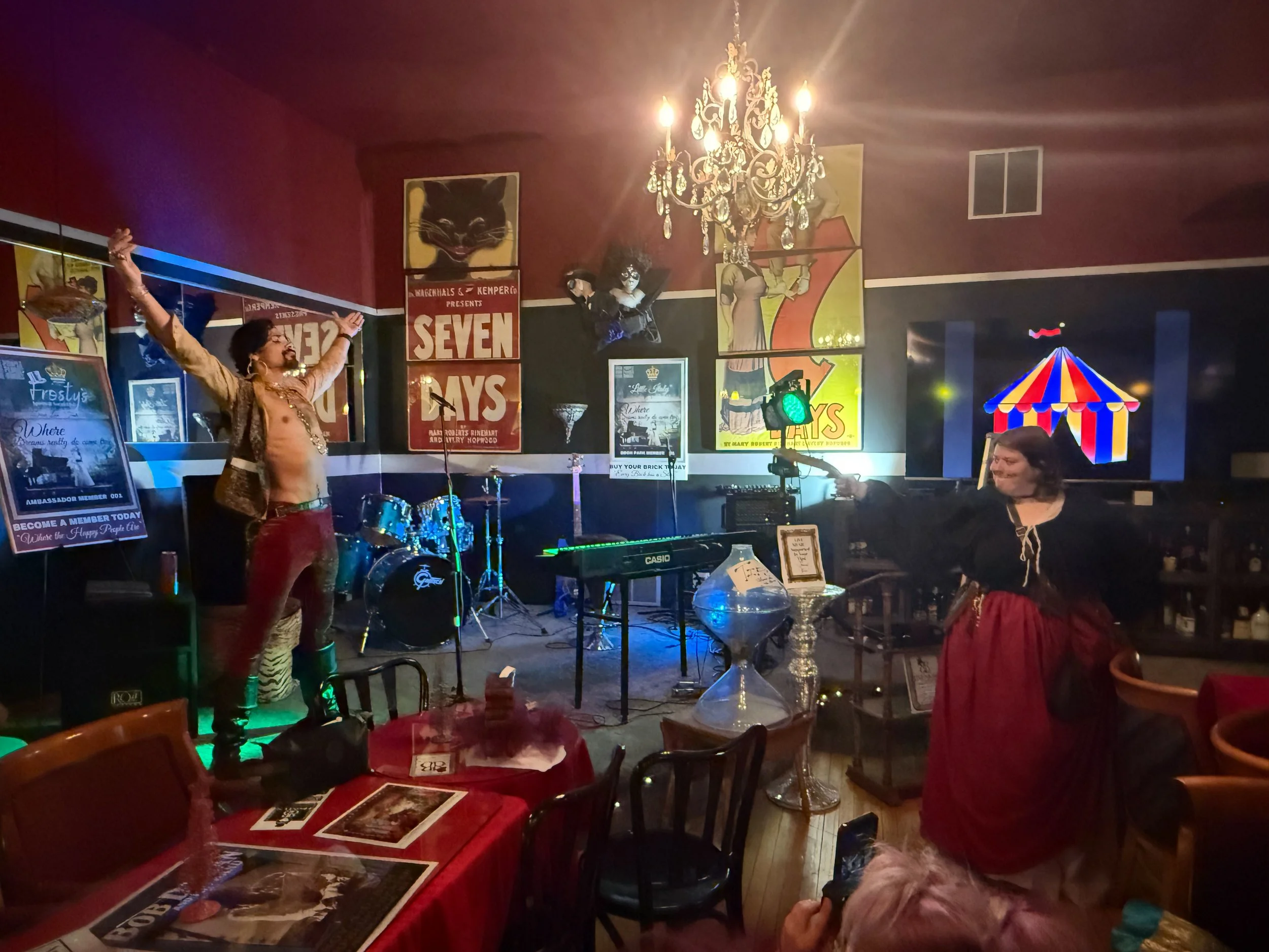 The sharp shooter and the escape artist are preforming their famous bullet catch at a lively bar or club with a stage, two performers dressed in vintage or theatrical costumes, musical instruments, and colorful posters on the wall. 