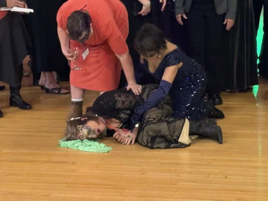People are helping a woman who appears to have fainted or fallen during a social event. She is lying on the floor with a green cloth under her head, and others are assisting her.