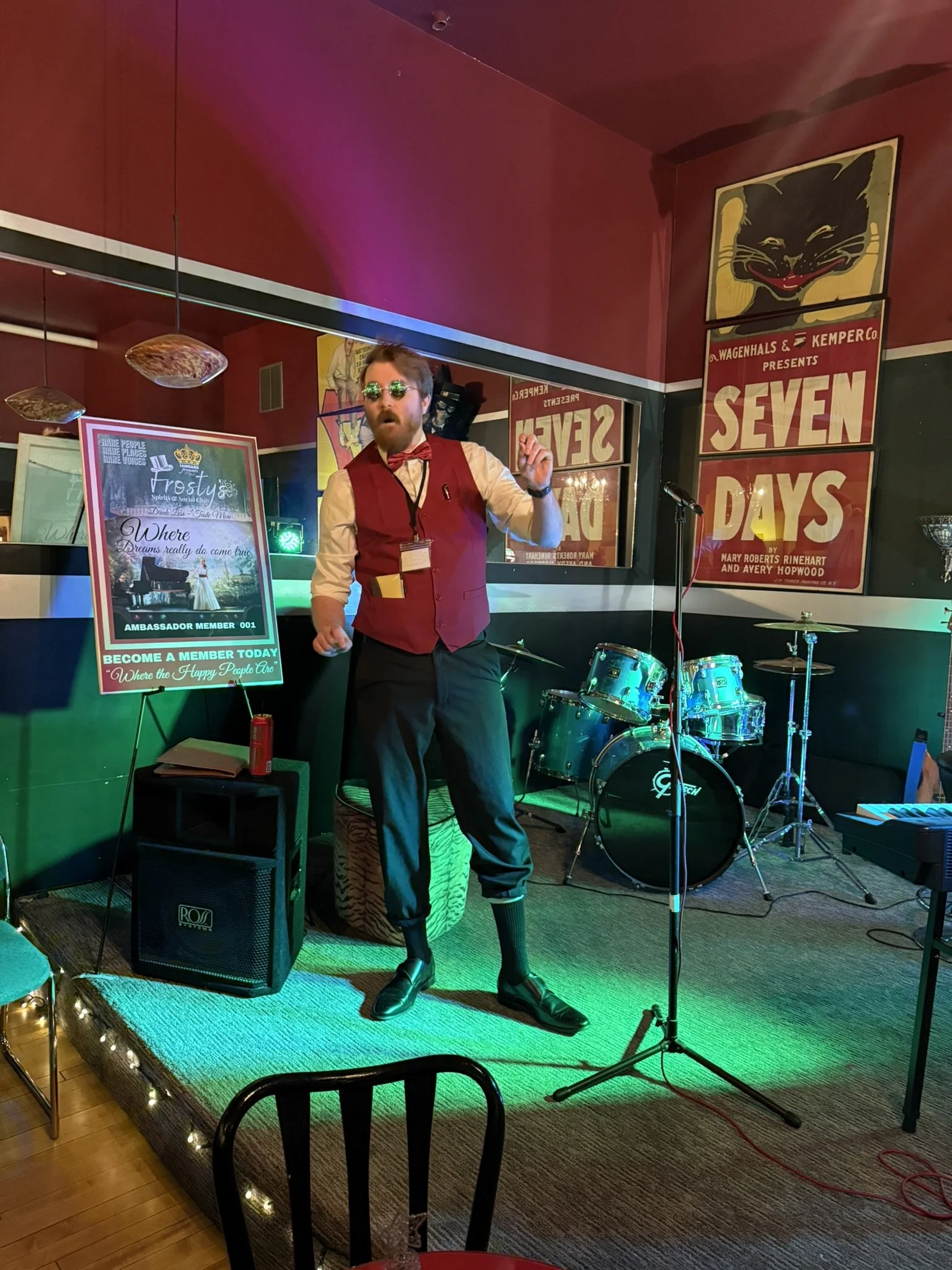 The carnival barker, with sunglasses and a red vest on a small stage in the New York Room, announces circus acts into a microphone, with posters on the wall behind him.