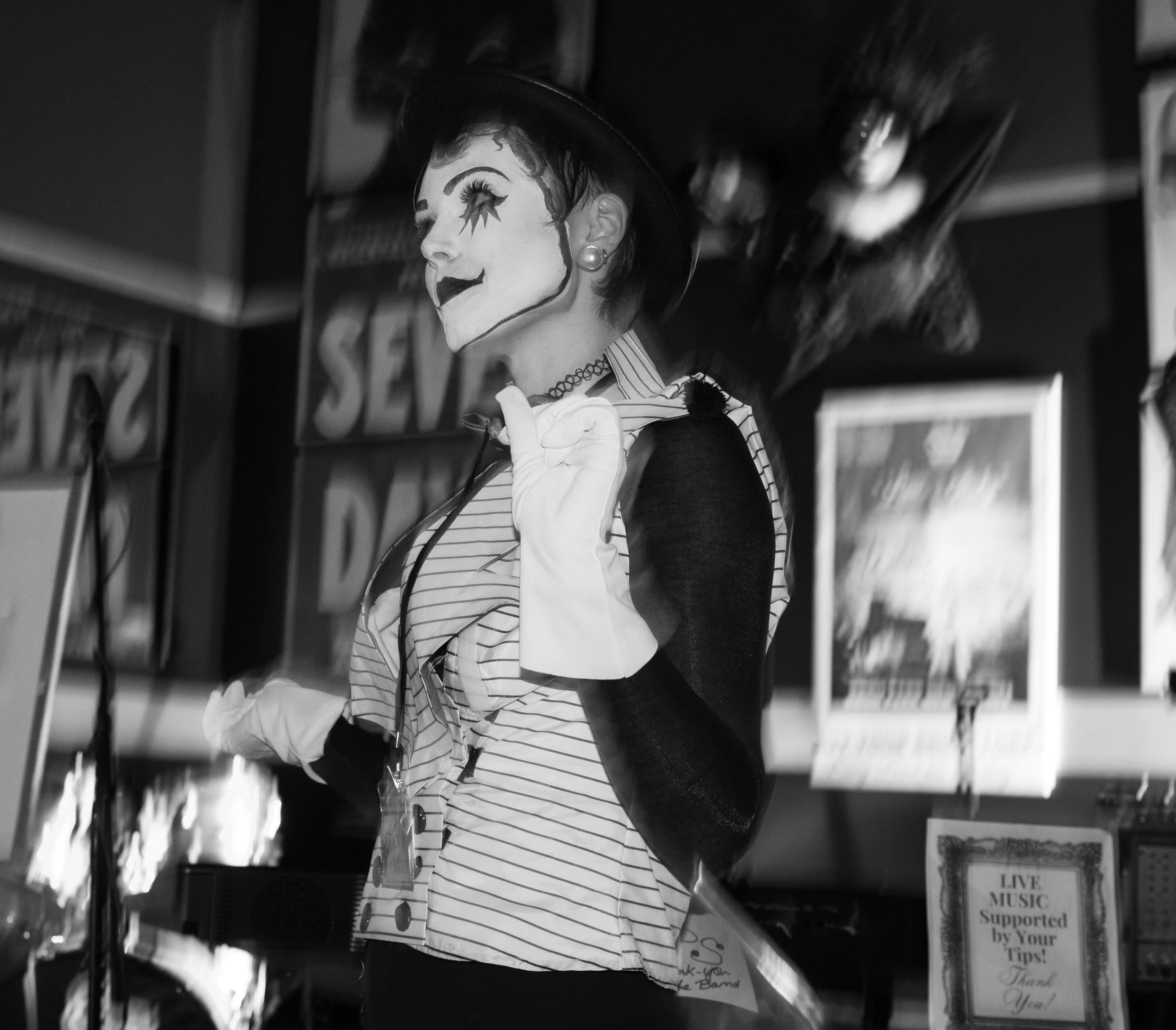 A greyscale image of woman dressed as a clown with mime makeup, wearing a striped shirt, black vest, and white gloves, on stage inside a venue with signs for live music at the Murder at the Circus Mystery Event
