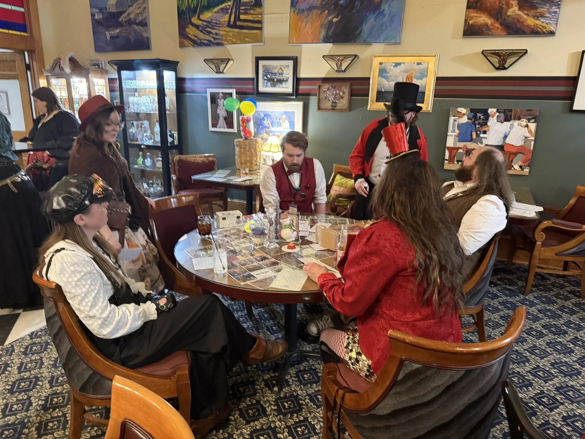 circus staff dressed in vintage circus and steampunk costumes gathered around a table in a decorated restaurant or cafe with paintings on the wall and balloons on the table.