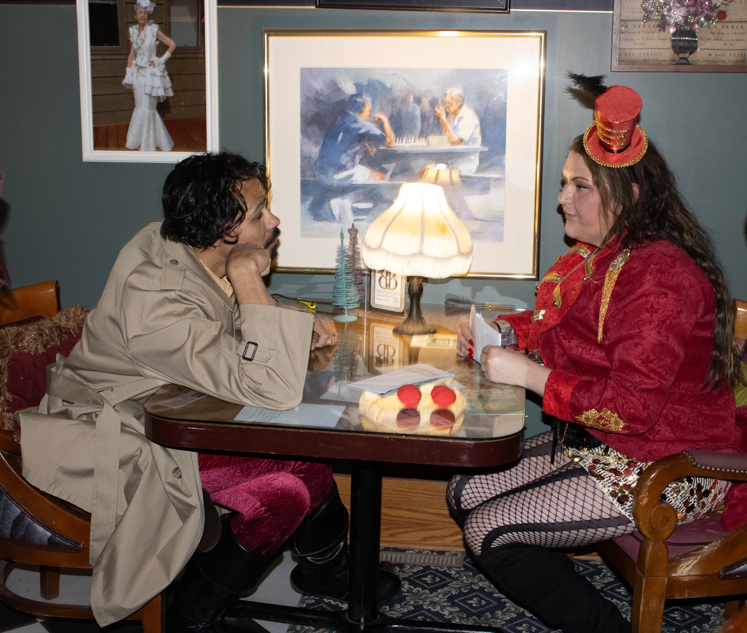 Lead Investigator Avery Clew, dressed in a beige trench coat and red velvet pants sits across from the ringmaster's assistant, in a red tailcoat with gold embellishments, fishnet stockings, and a small red top hat with a feather, in a cozy room decor