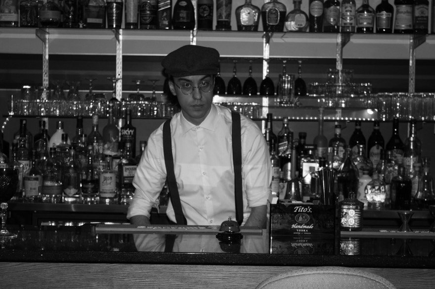 A grayscale image of a bartender with glasses, wearing a white shirt, suspenders, and a beret, standing behind a bar with various bottles of alcohol on shelves.