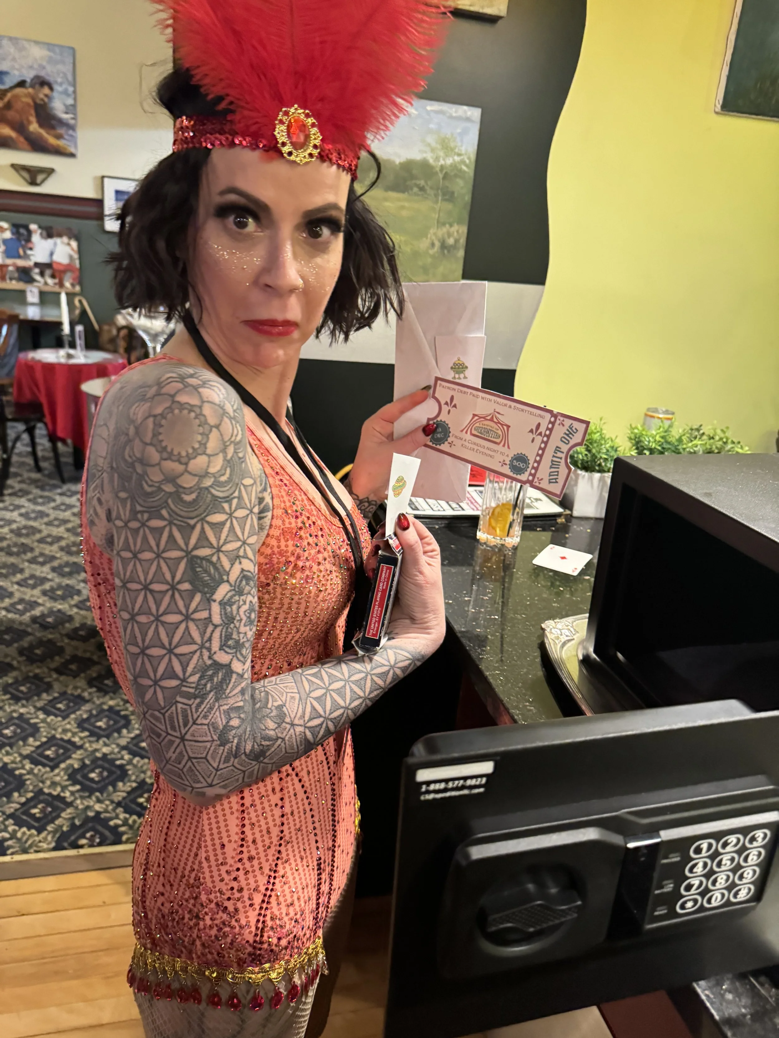 A woman dressed as a trapeze artist in a colorful, sequined outfit with a large red feathered headpiece and tattoos on her arms, standing at an unlocked safe, looking surprised while holding a circus entry ticket.