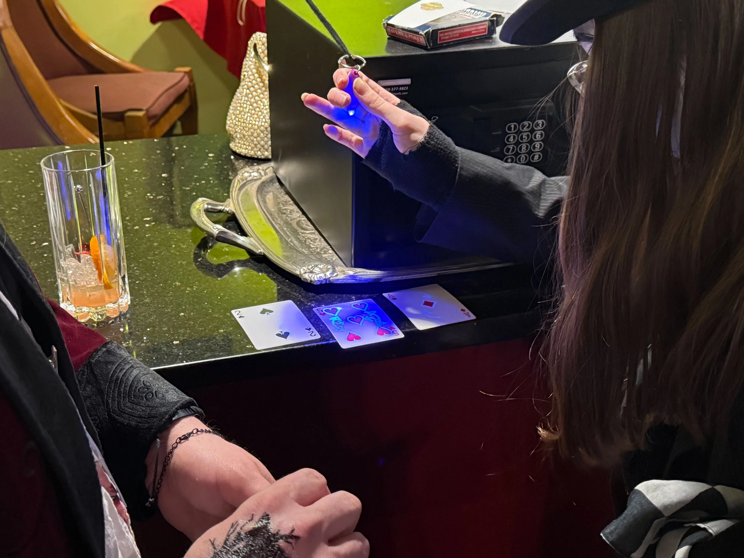 A secret message written on playing cards is revealed under blacklight. The puzzles rests on a bar, with a glass of drink, a decorated bag, a plate with a fork, and a safe in the background.