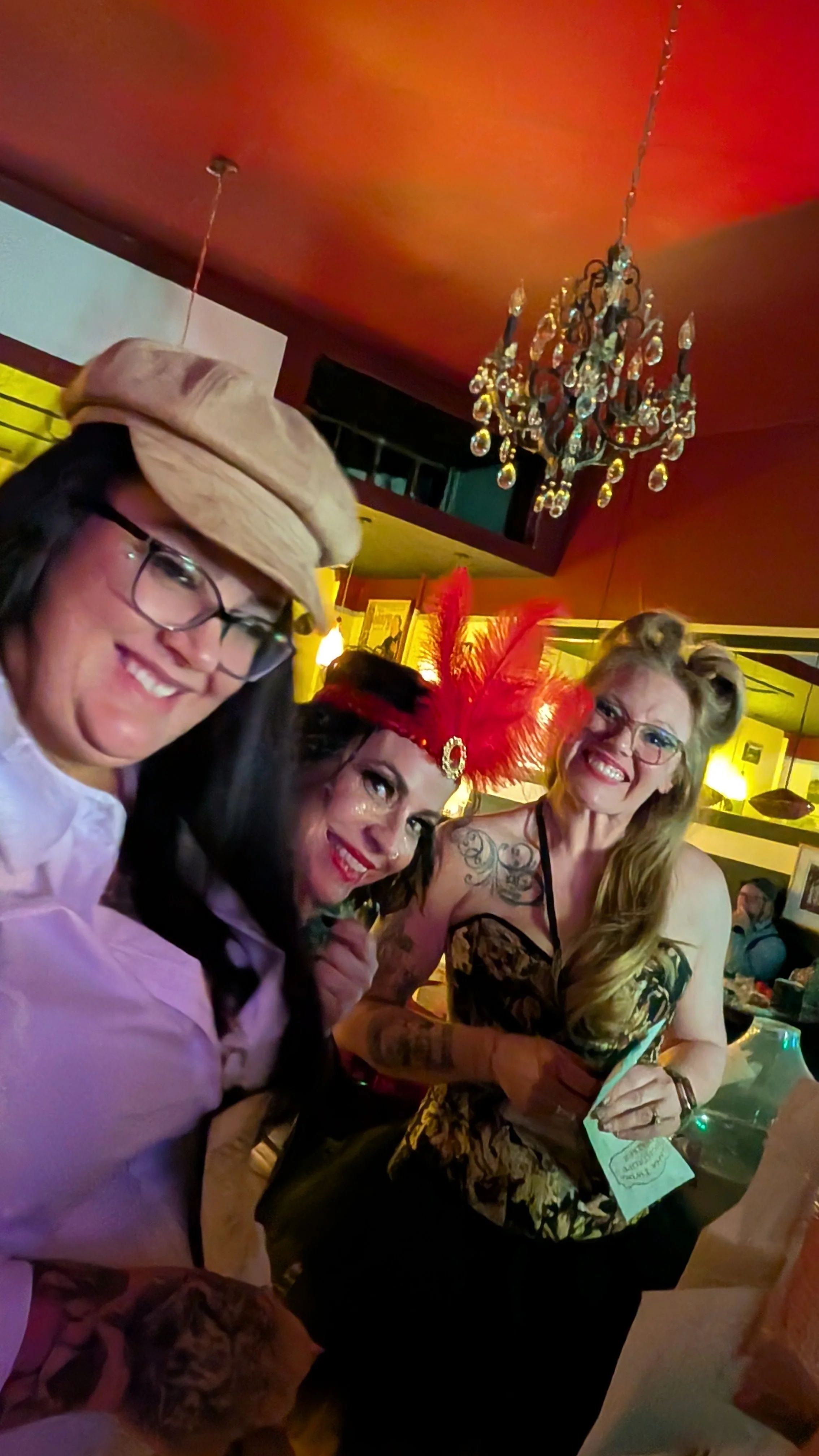 A detective, trapeze artist, and tightrope walker at a festive event, with a chandelier hanging from the ceiling, one wearing a beige hat, another with red feathers on her head, and the third with glasses and a tattoo on her chest, smiling and holdin
