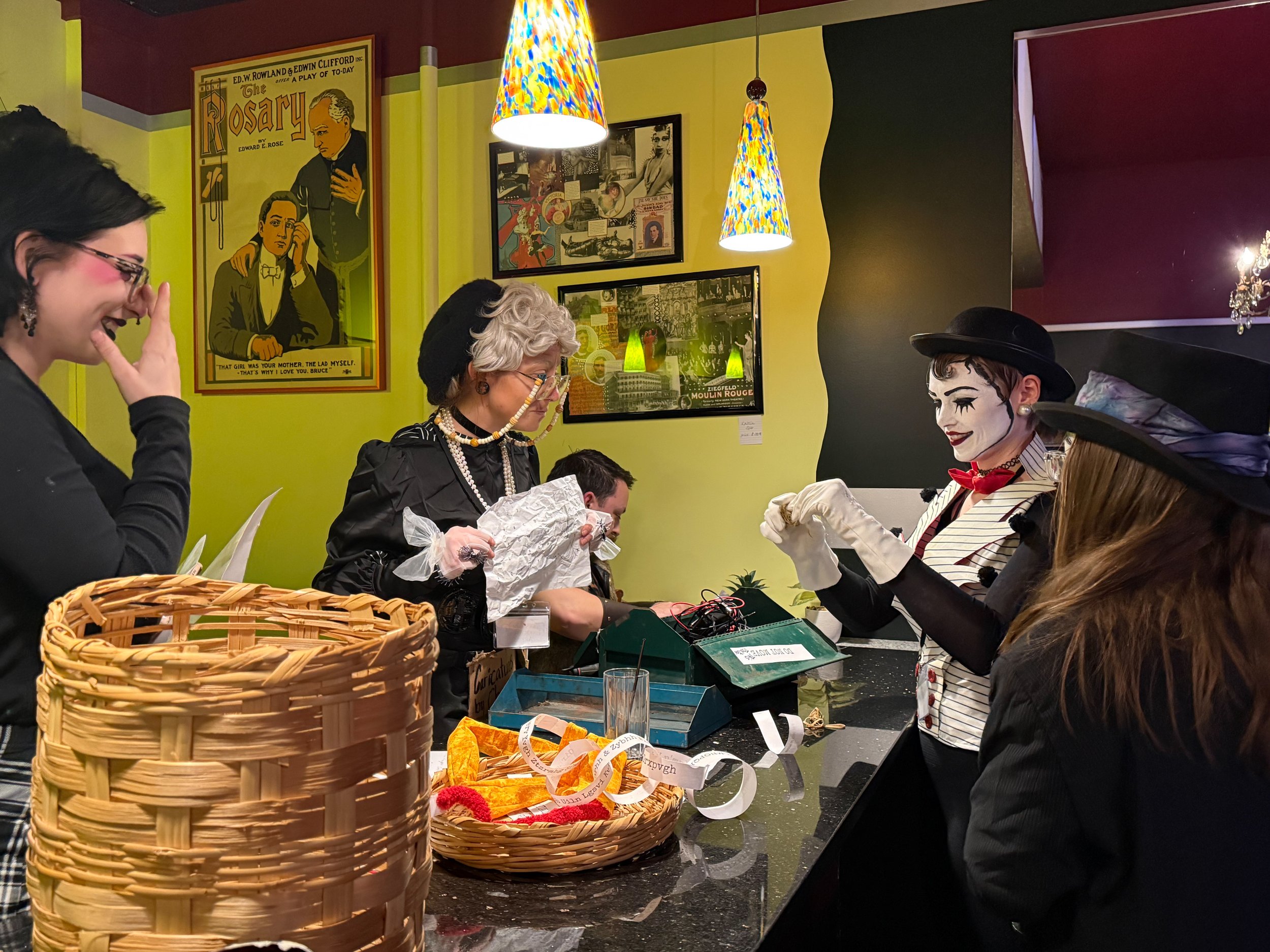 The contortionist, mime, caricature artist, and tramp clown, all dressed in costume, gather around a counter  full of puzzles in a colorful, retro-themed room decorated with framed posters and bright hanging lamps. One puzzle consists of a toolbox, n