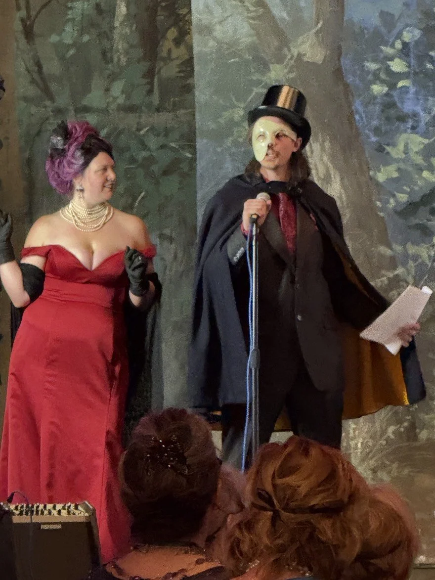 The Murder Mystryss and the Phantom stand on a stage, her in a red evening gown with pearls and gloves, the him in a black cape, top hat, and a half mask holding a microphone, with audience members in the foreground.