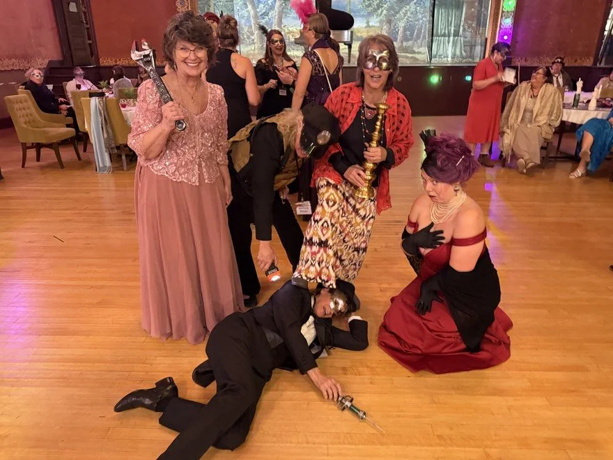 Murder Mystery party guests in elegant costume posing in a ballroom with faux murder weapons. One guest laying on the floor as the assumed victim. The Murder Mystryss kneeling at the scene.