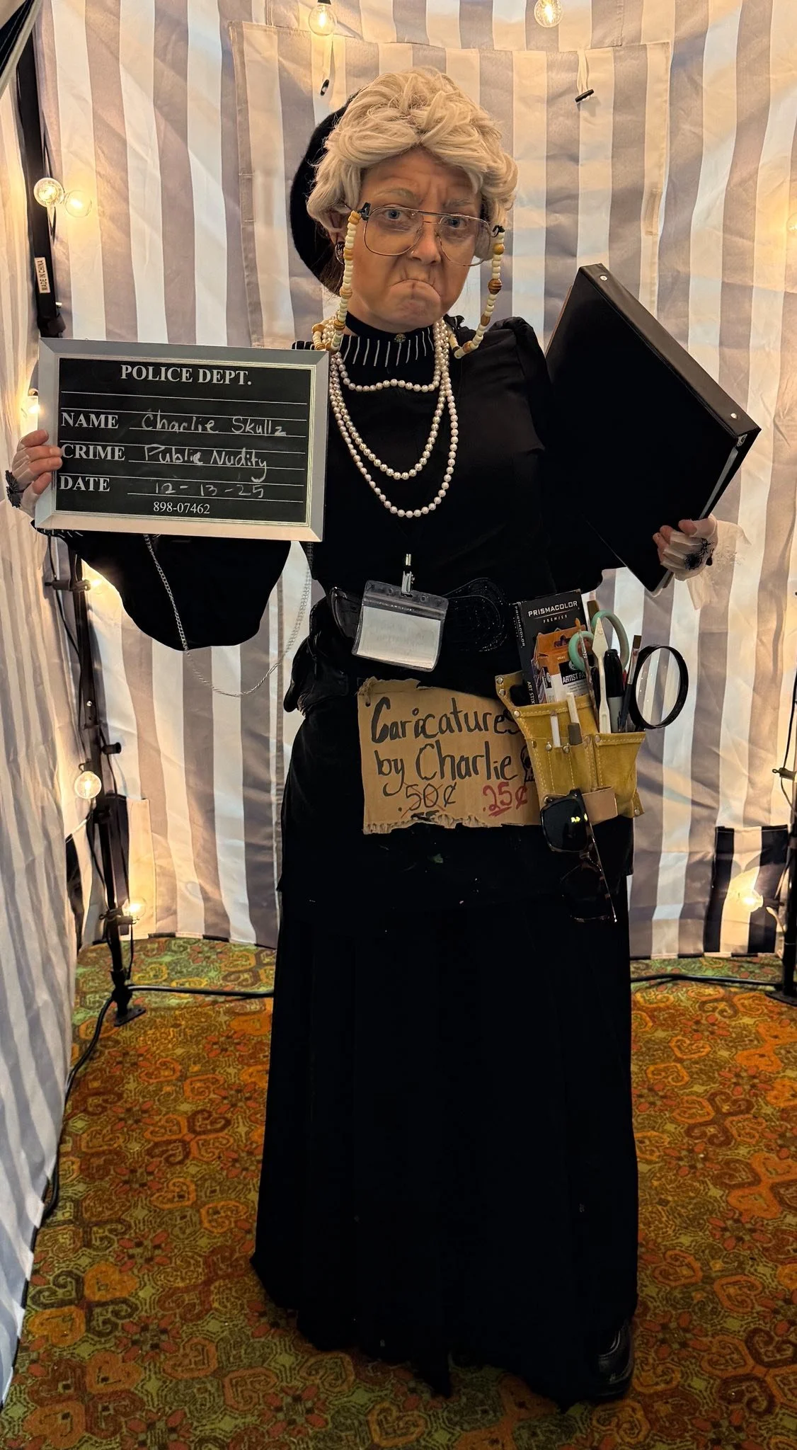 Person dressed as an old woman, holding a police sign and a folder, with a sign on her waist advertising caricatures by Charlie for 50 cents. She wears glasses, a wig, and pearl necklaces, and has a stern expression. The background is striped fabric 