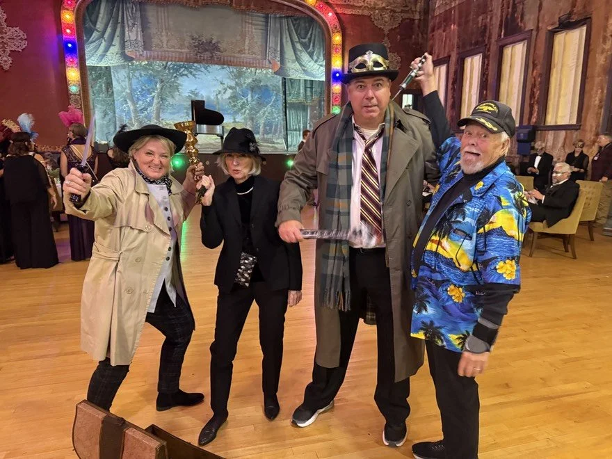 Four people dressed in detective costumes and accessories, posing for a photo at a masquerade party or event with a colorful, illuminated stage in the background.