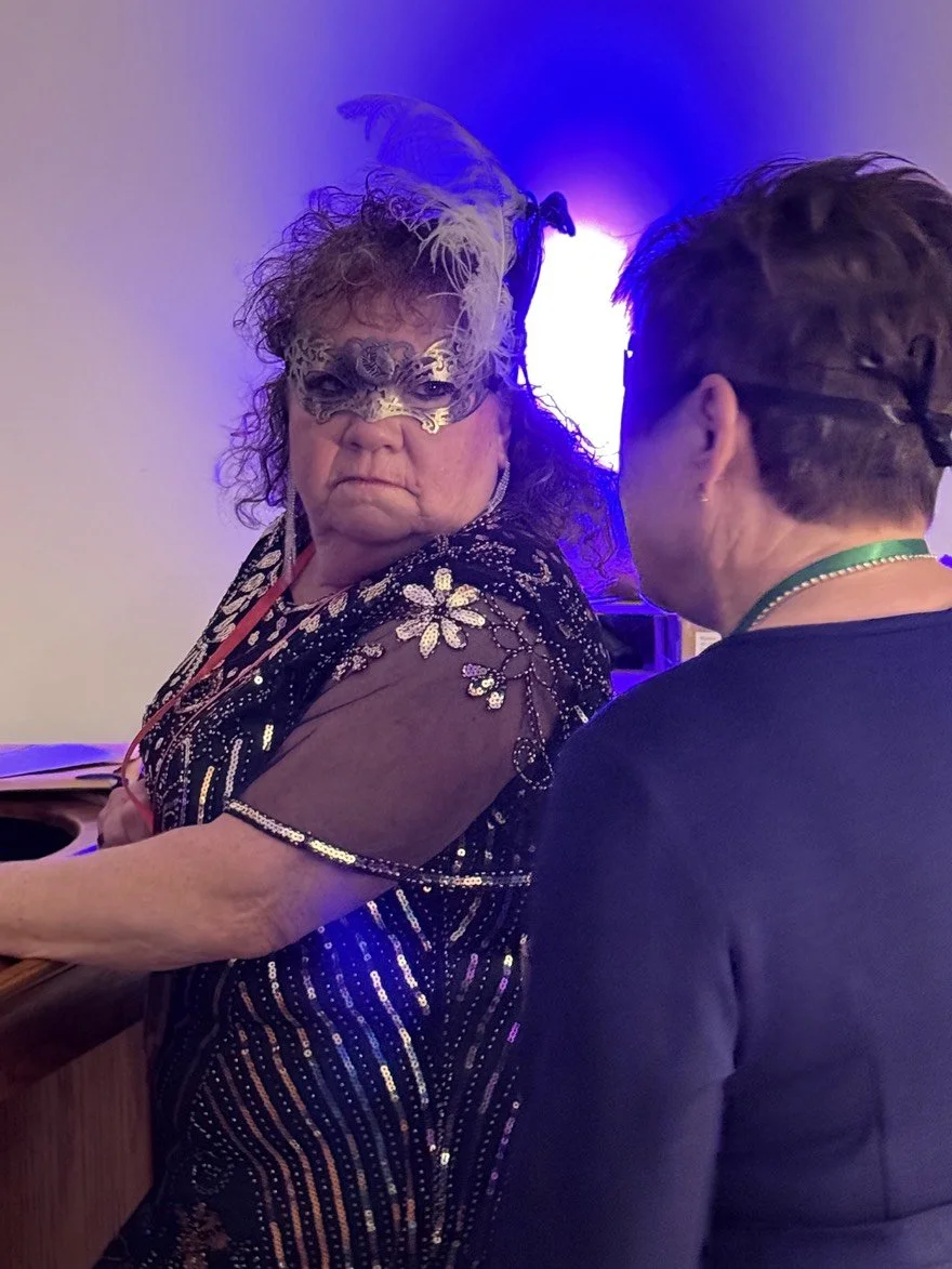 Lola Delacroix, the Showgirl, looks as if she is defending against being accused. She is engaged in conversation at an indoor event. She is dressed in an elaborate, sequined dress with a feathered mask and feathered headpiece, speaking to another in 
