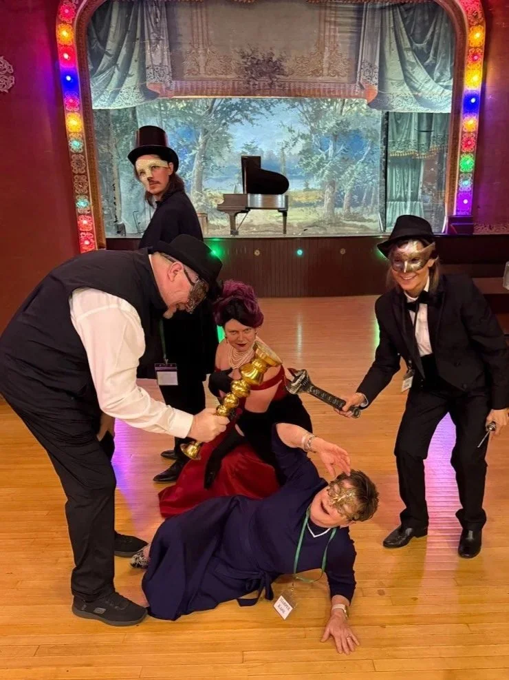 Murder Mystery party guests dressed in costumes, wearing masks and holding fake weapon props, pose as suspect and victim in front of a stage with a backdrop of a park scene and a piano in the background. The Murder Mystryss smirks at the scene.