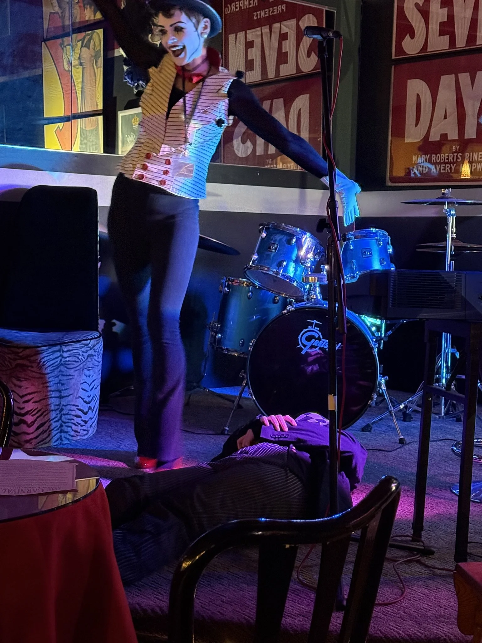 A performer dressed as a clown or mime with a painted face, wearing a striped vest, black shirt, and black pants, standing on a small stage with a drum set in the background. The performer is smiling and appears to be in a lively moment, with colorfu