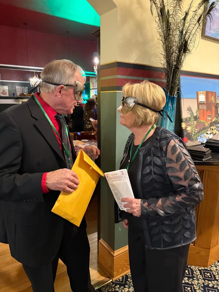 Two older adults, a man and a woman, wearing masks, are deeply engaged in conversation at a social event in a restaurant or banquet hall. The man is dressed in a black suit with a red shirt, holding a yellow envelope, and the woman in a patterned bla