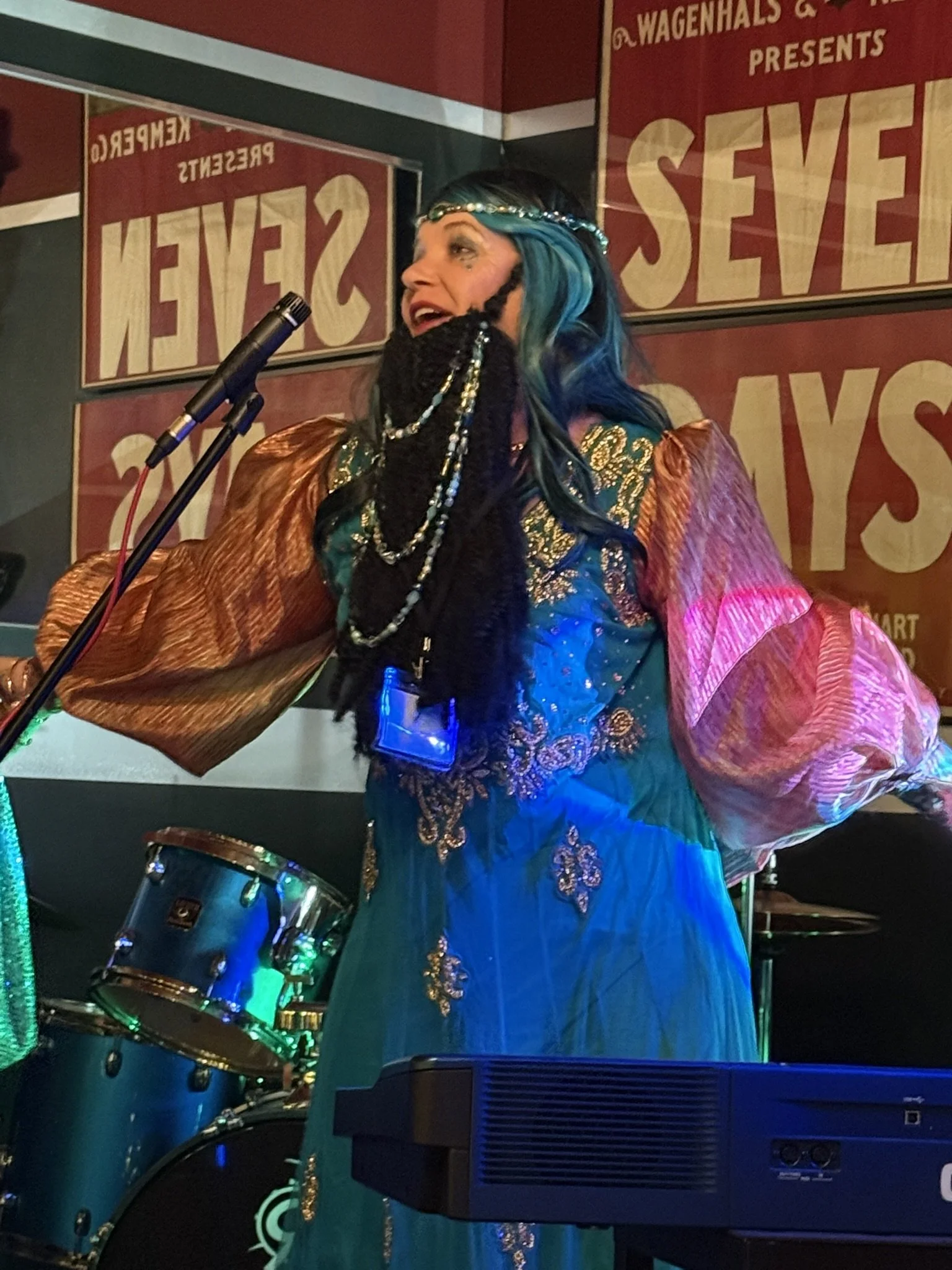 The bearded lady, with blue hair, wearing a jeweled headband, black gloves, and a colorful, ornate dress with gold embroidery, standing at a microphone on stage. She is smiling and appears to be singing or speaking. Behind her are posters and musical