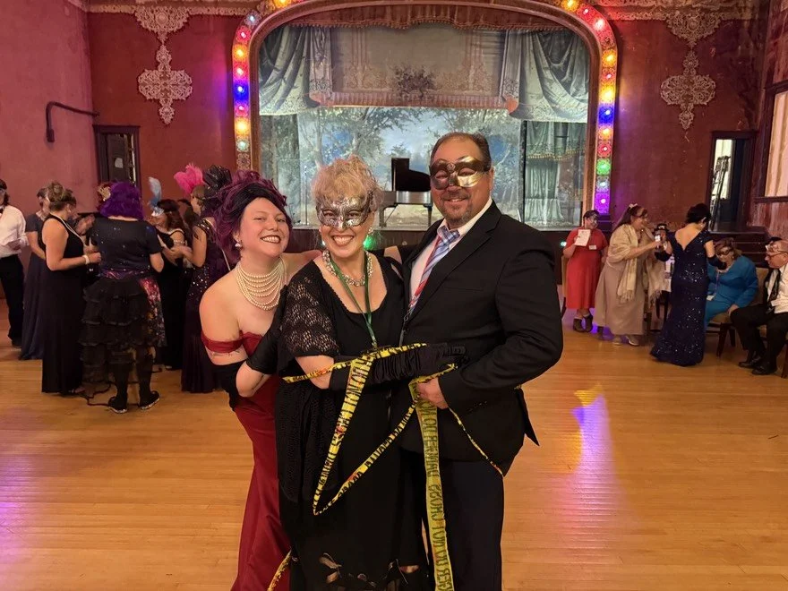 The Murder Mystryss has wrapped a couple in crime scene tape. Both are dressed in elegant costumes at a lively masquerade party or event in a decorated ballroom with colorful lights.