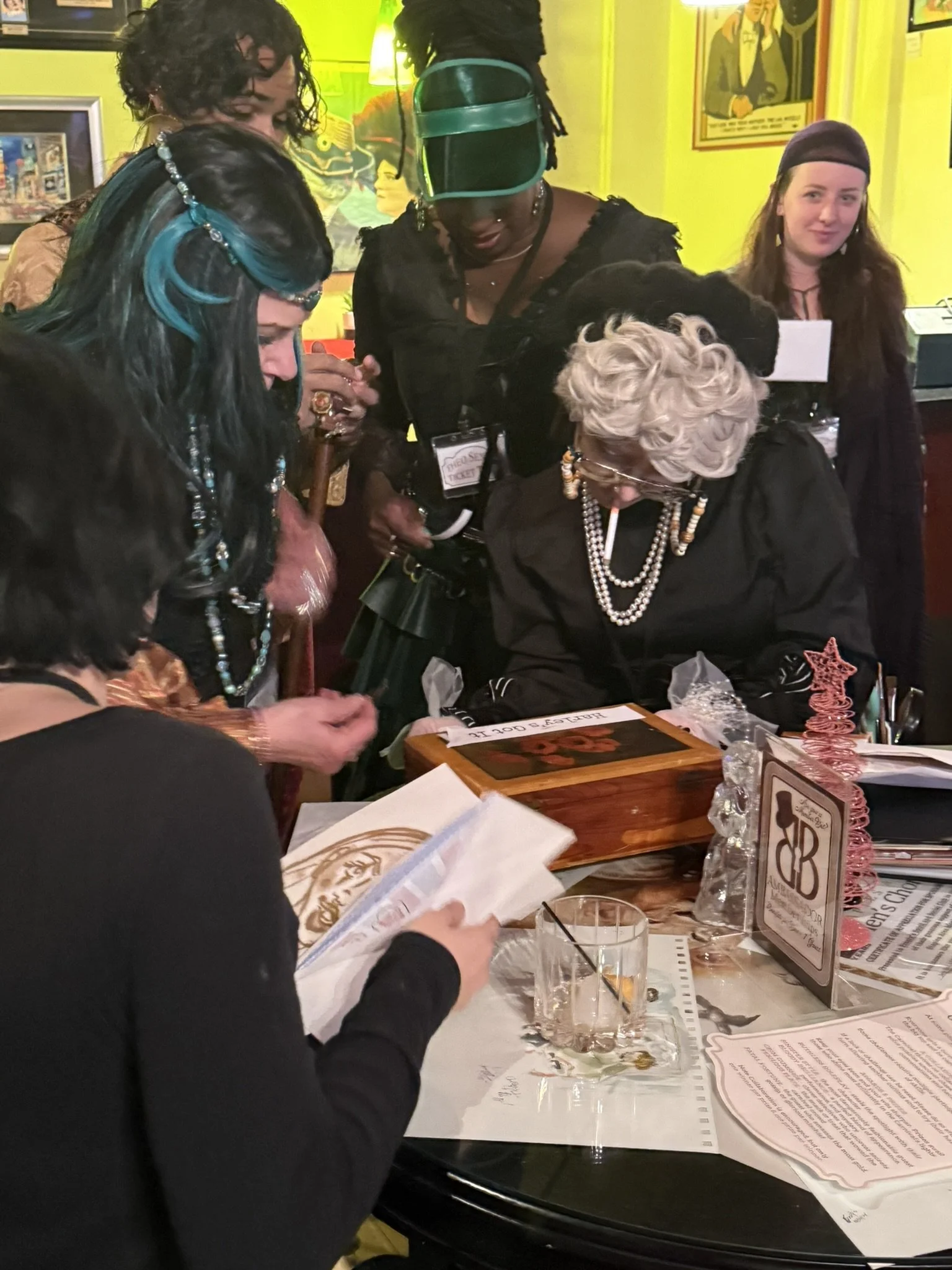 A group of circus performers dressed in stylish, eclectic clothing and accessories gathered around a table in a colorful, retro-themed restaurant or bar, looking at puzzles and clues