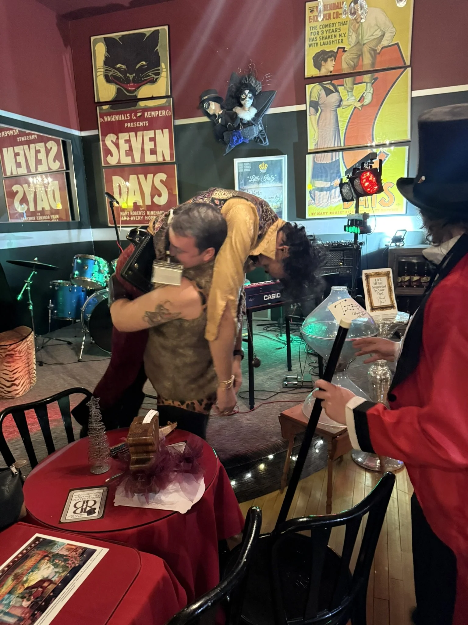 People dancing in a lively room with circus-themed and vintage posters on the wall, including a black cat, seven days, and a man and woman in vintage clothes. There is a drum set in the background, a red table with a small decorative tree and a wrapp
