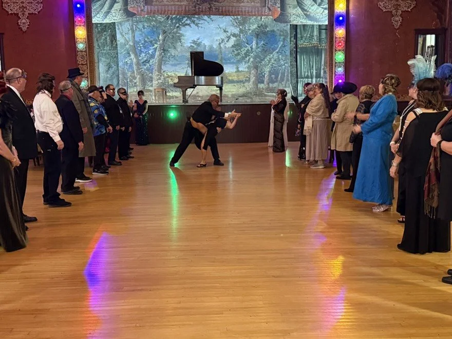 People dressed in vintage attire are gathered in a ballroom, watching a couple perform a dance on a wooden floor. He is dipping her gracefully. A grand piano is visible on a stage with a scenic backdrop, and colorful lights decorate the room.