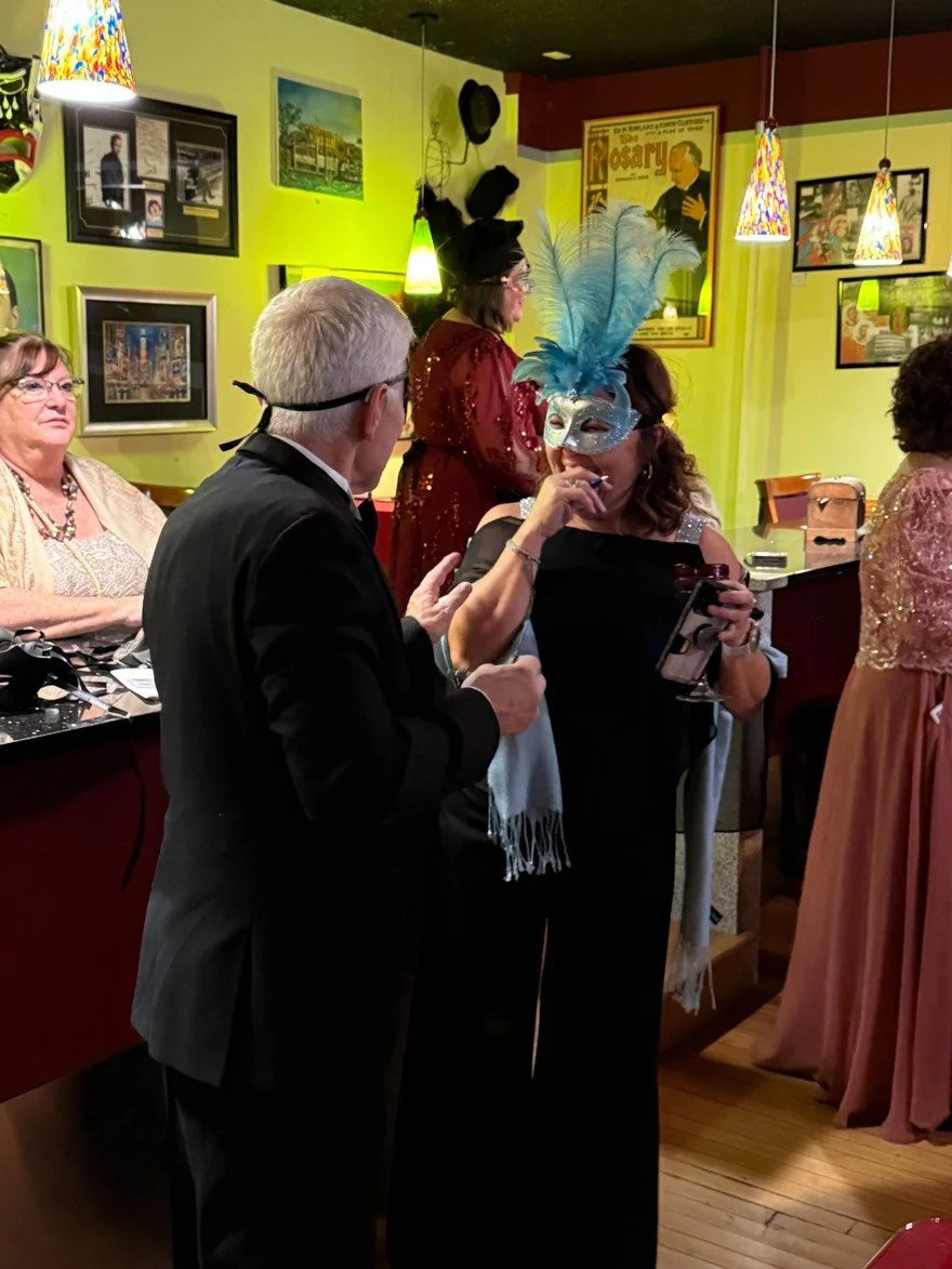 Two guests of the Phantom of the Batcher Block Opera House Masquerade Ball laugh in the New York Room. The woman is wearing an elegant gown with a tall blue feathered mask. The man wears a suit and mask,