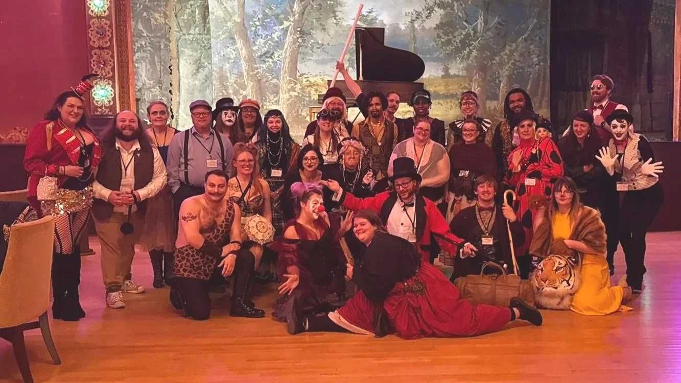 Murder at the Circus group photo. The cast is dressed as various carnival performers and staff. All posing together in front of an opera house stage. The Murder Mystryss kneels in front dressed as a burlesque clown while the sharpshooter does splits