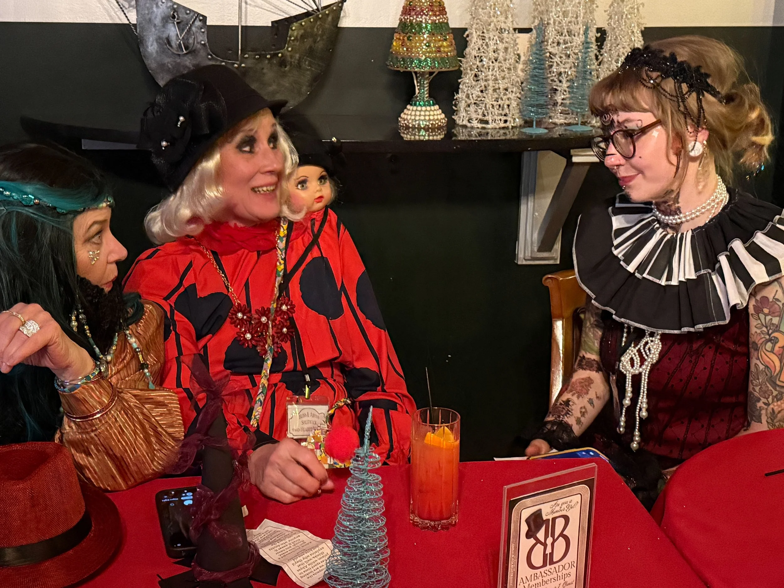 The bearded lady, two-headed wonder, and the most tattooed person at a festive gathering dressed in colorful, eclectic costumes, with a clue and a drink on the table.
