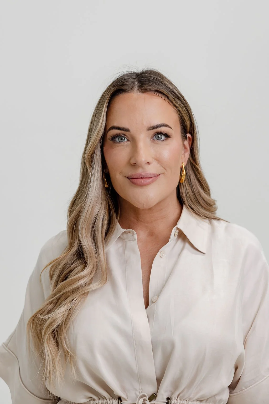 A woman with long wavy blonde hair, blue eyes, wearing earrings and a cream-colored button-up shirt, smiling at the camera against a plain white background.