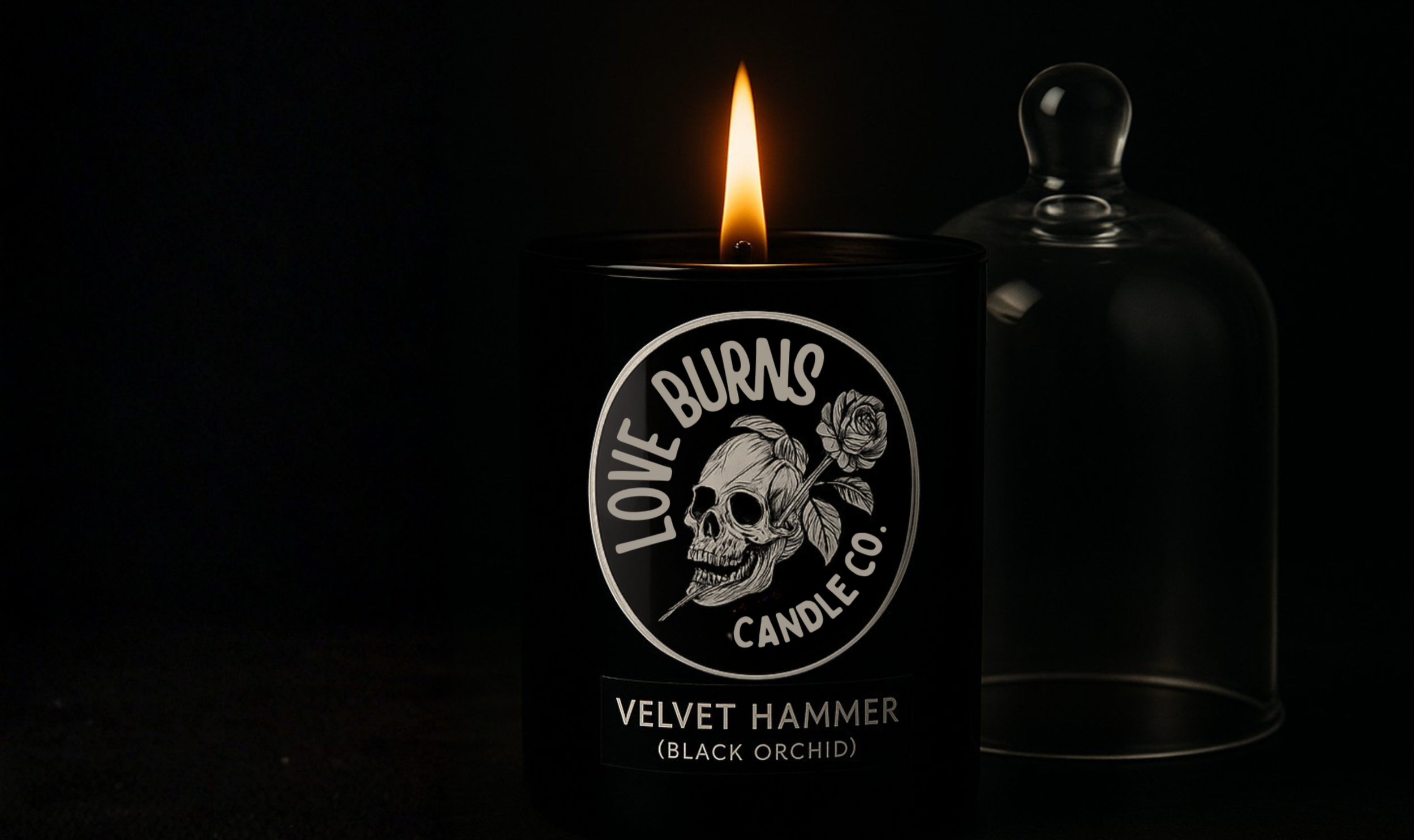 A black candle with a skull and rose graphic on the label, labeled 'Love Burns Candle Co. Velvet Hammer (Black Orchid)', burning in a dark setting with the flame visible. A glass cloche is beside it.