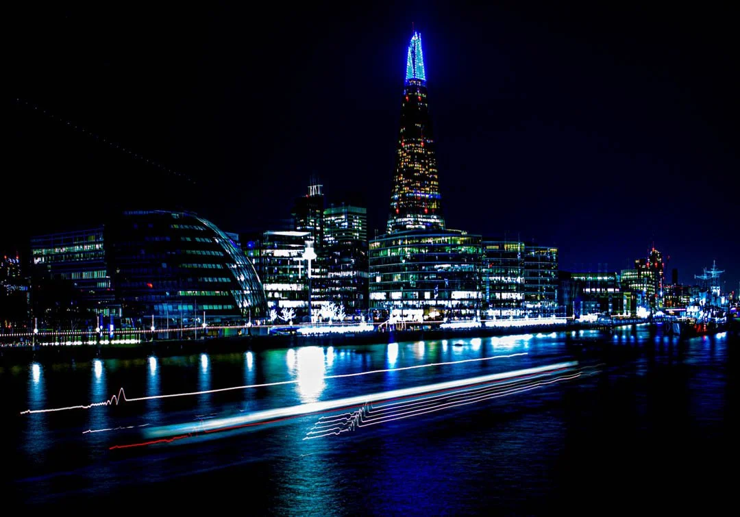 Night view of London skyline with illuminated buildings and the Shard skyscraper reflected on the river, with boat lights creating a pulse on the water.