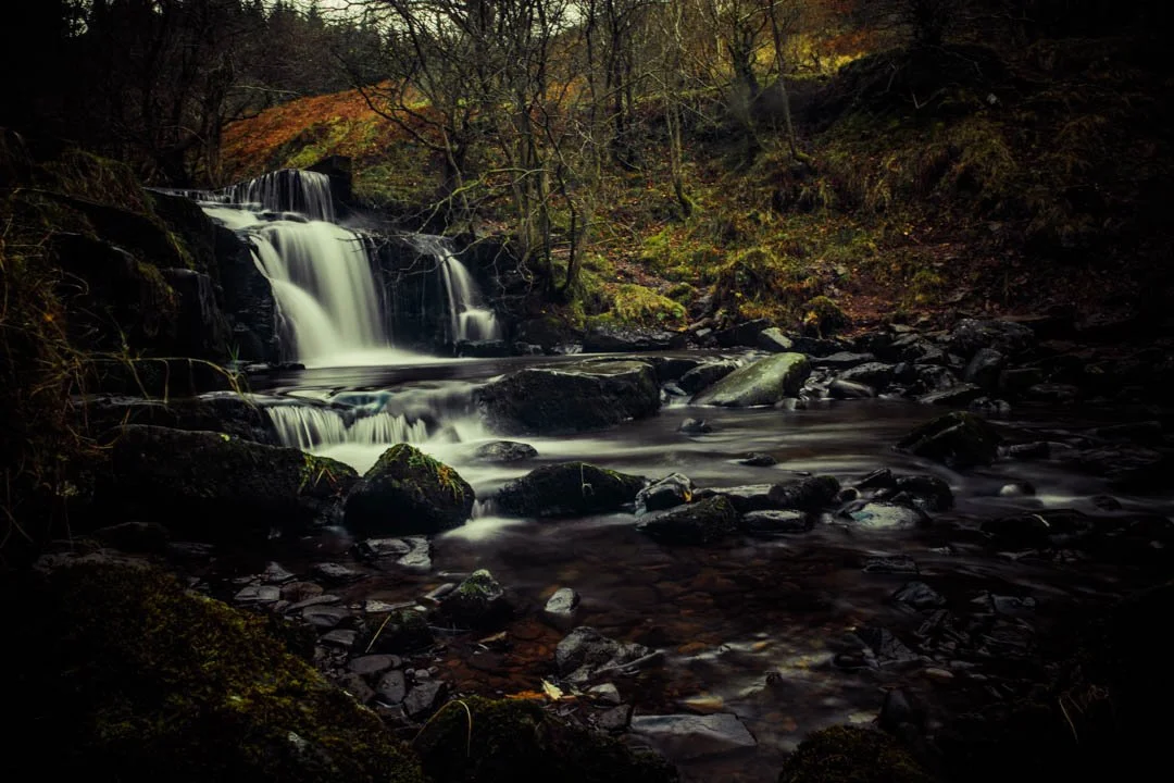 A small waterfall flows over rocks in a forested area during autumn.