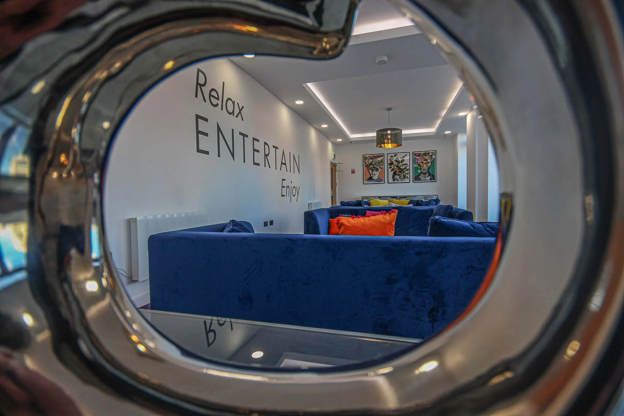 A modern lounge with blue velvet sofas, colorful pillows, and wall art of floral portraits, as viewed through a metallic circular frame.