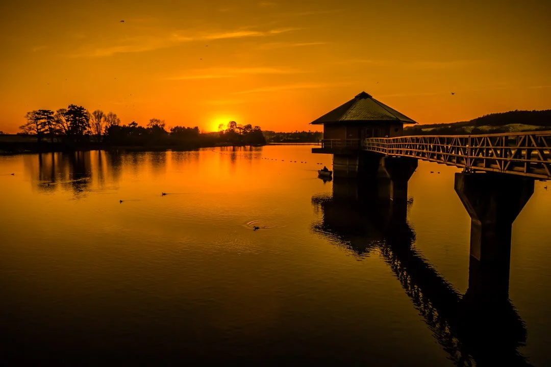 Sunset over a calm lake with trees on the horizon and a small house on stilts connected by a wooden bridge.