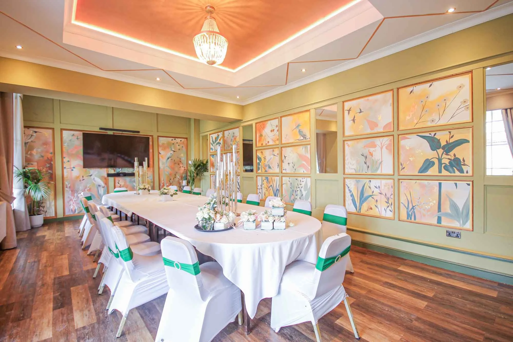 Elegant dining room with a long oval table set with flowers and candles, surrounded by white chairs with green sashes, decorated with pastel-colored bird and floral wall art, wooden floor, and a chandelier overhead.