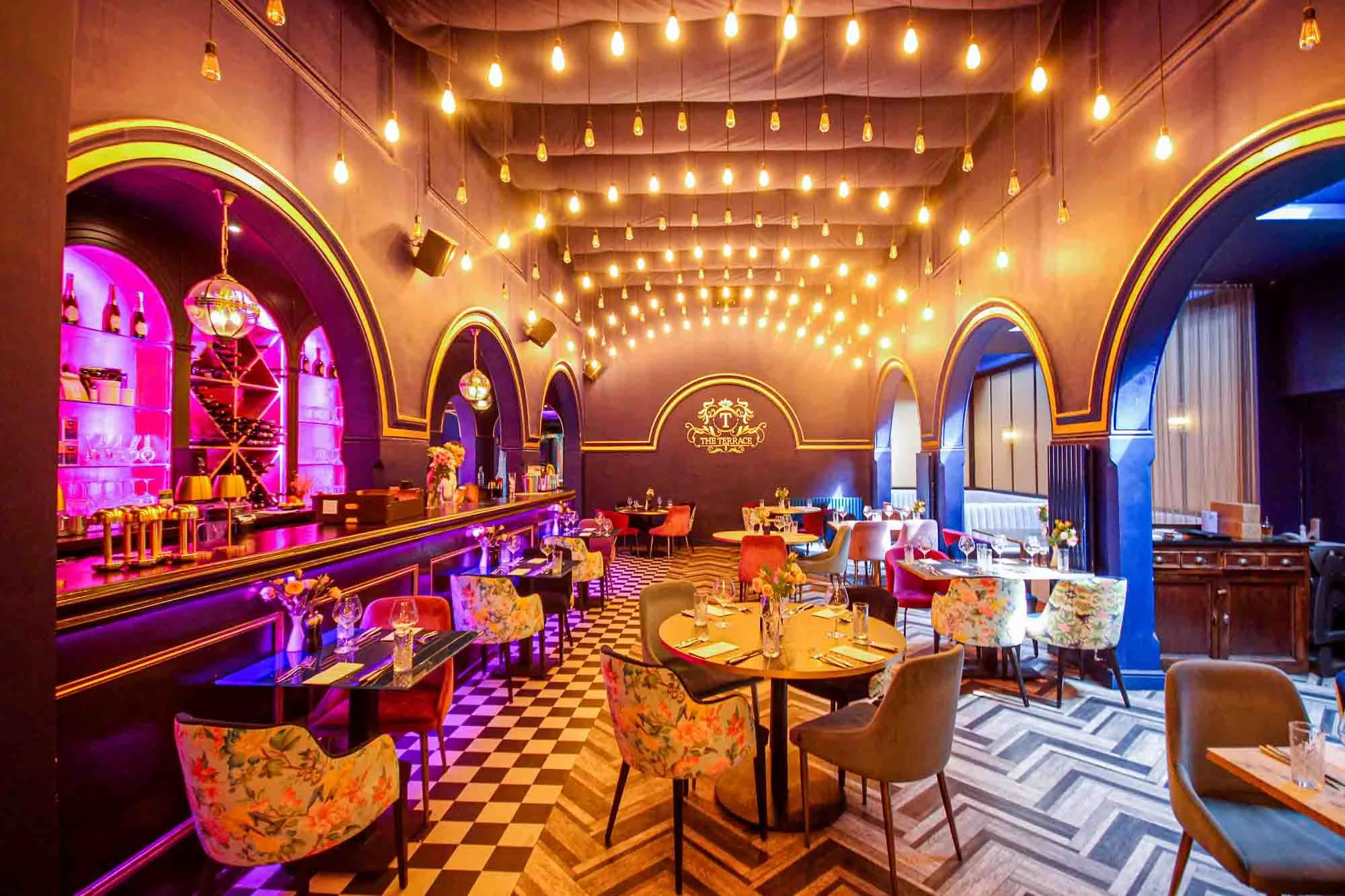 A colorful, stylish restaurant with purple and pink lighting, arranged with round tables set with glasses and cutlery, floral chairs, and a checkered floor. The ceiling has string lights, and a bar area is visible with bottles and glasses, alongside 
