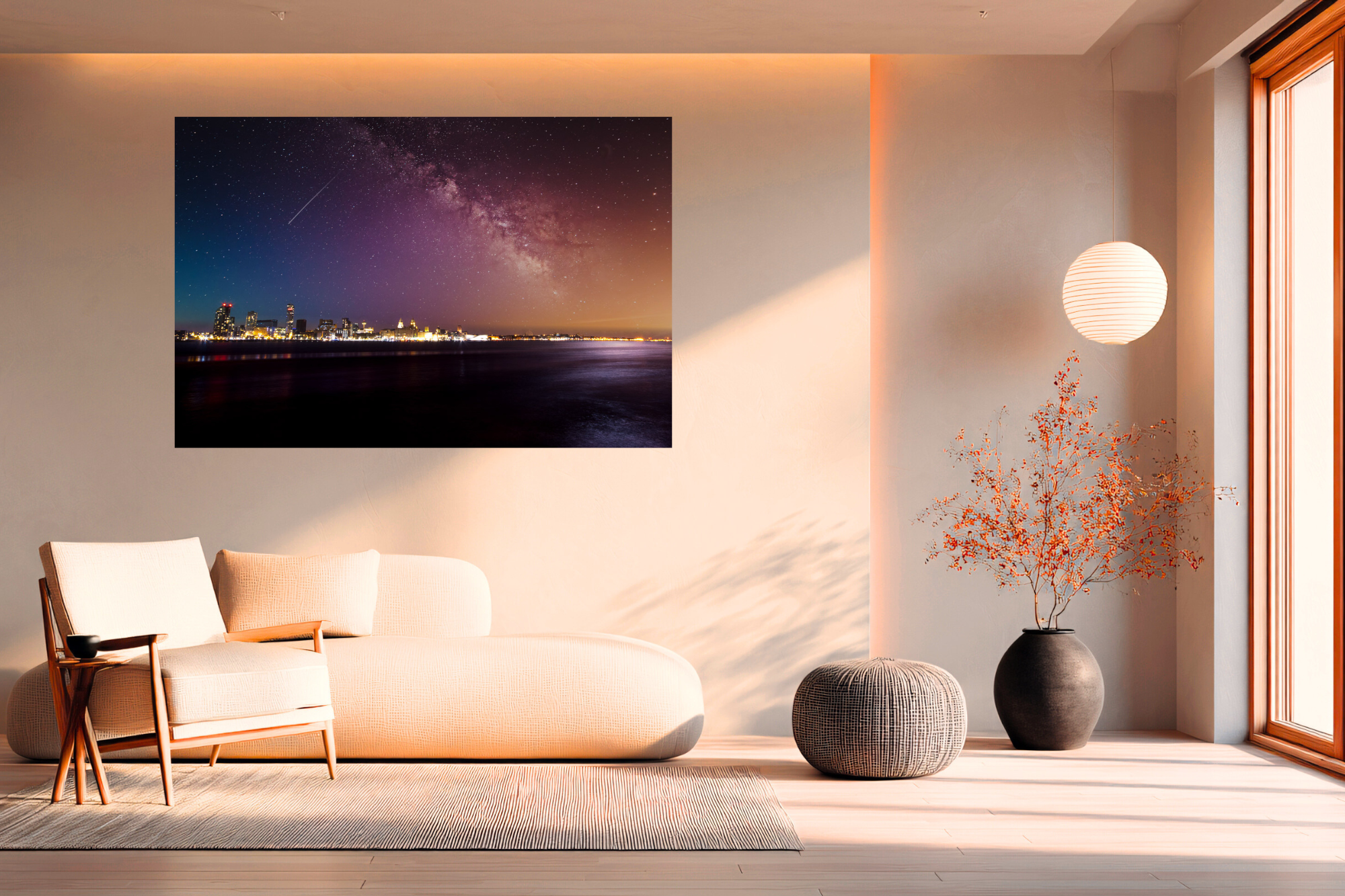 Modern living room with a beige sofa, a black vase with orange foliage, a hanging paper lantern, and a large landscape photograph of a night city skyline with the Milky Way in the sky, sunlight streaming through a window.