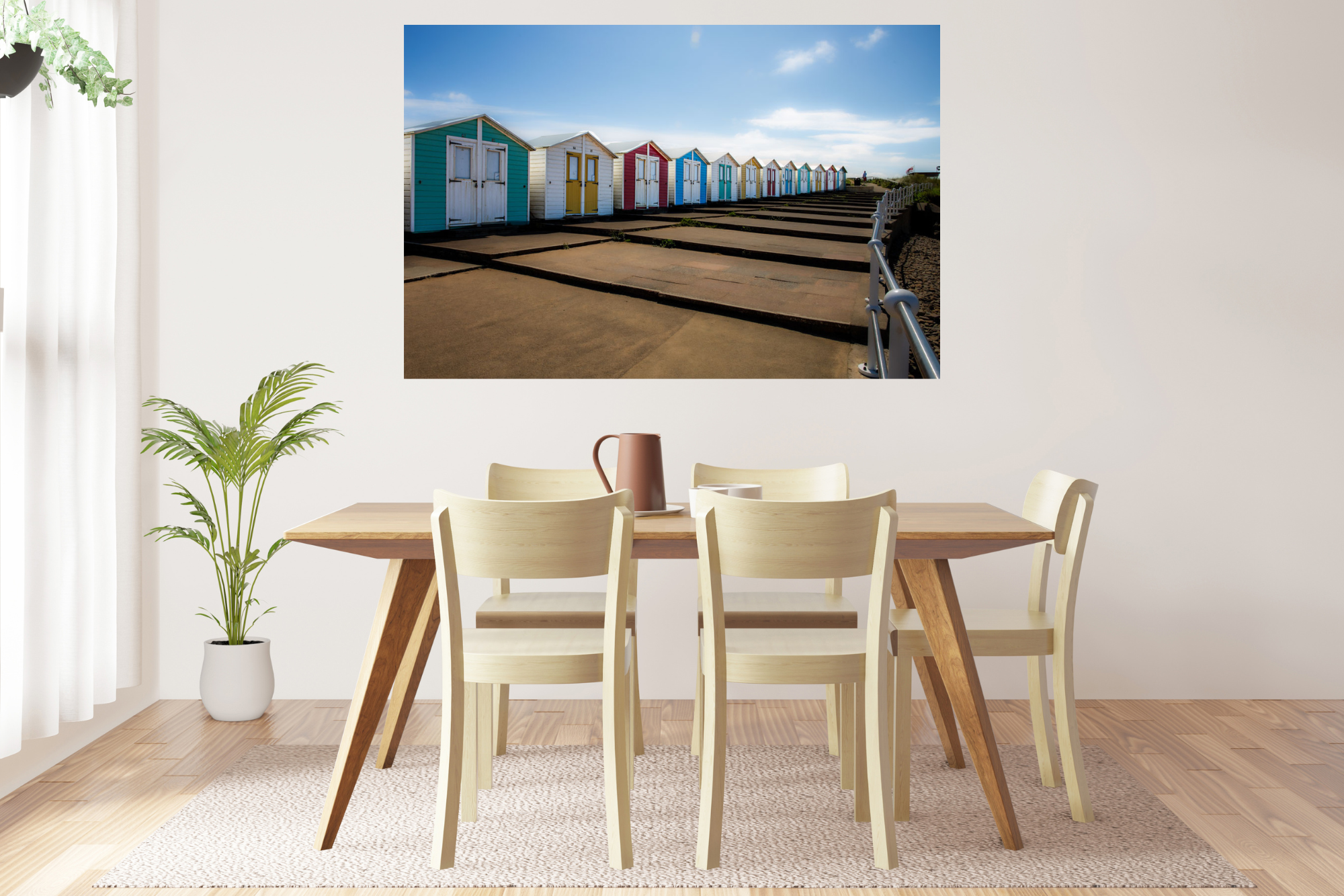 A photo of colorful beach huts with a walkway and sky above, hanging on a white wall in a dining room with a wooden table, four white chairs, a potted plant, and a vase on the table.
