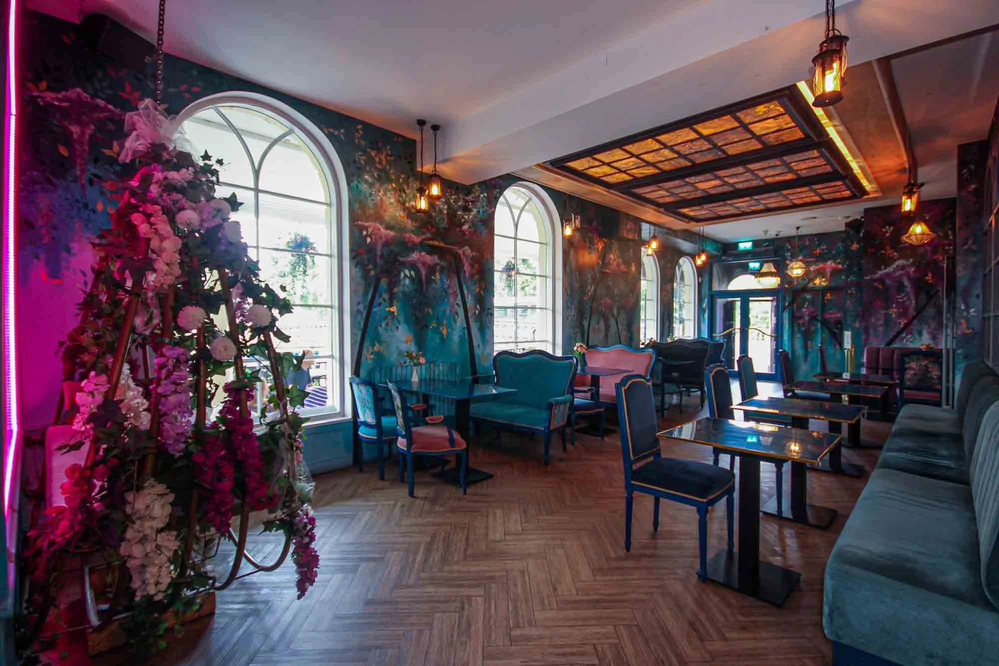 Interior of a decorated cafe or lounge with arched windows, colorful floral wall murals, vintage-style chairs, tables, and hanging lamps, creating a vibrant and artistic atmosphere.