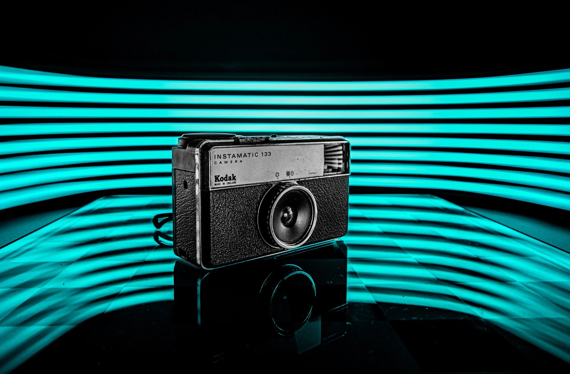 Vintage Kodak Instamatic 133 camera placed on a reflective black surface with glowing blue neon light strips in the background.